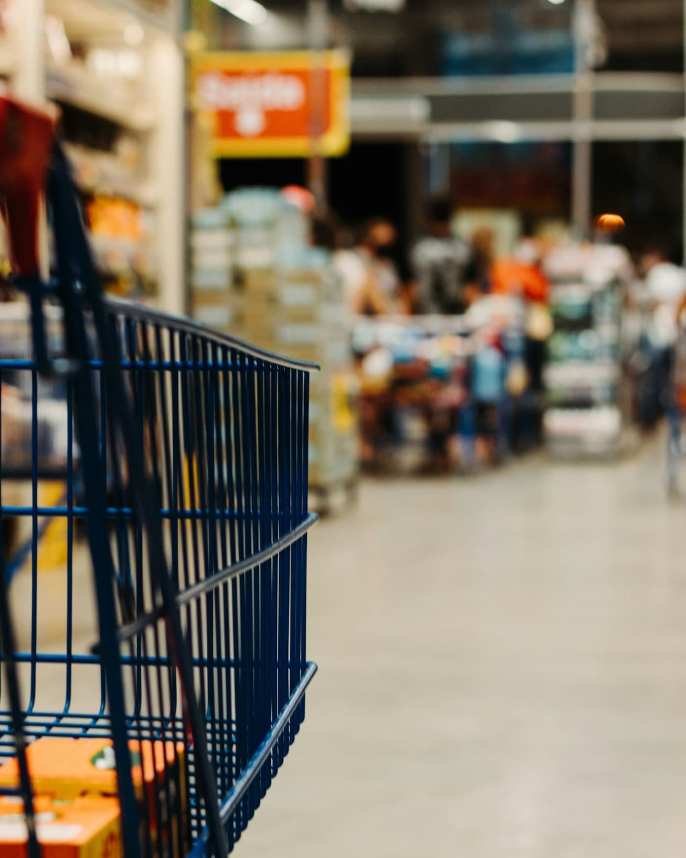 Blue shopping cart in a busy warehouse-store aisle, shoppers out of focus